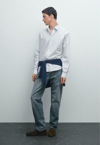 Camicia bianca a righe con bottoni, abbinata a jeans blu e mocassini marroni scuri; maglione blu navy annodato in vita.