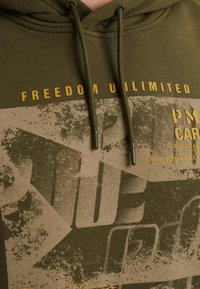 Donkergroene hoodie met gele tekst en grafisch ontwerp met een textuurpatroon. Verstelbare trekkoorden met metalen punten. Casual pasvorm.