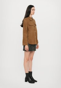 Giacca camicia in suede marrone con chiusura a bottoni e due tasche frontali, indossata sopra una minigonna nera e abbinata a stivaletti neri con tacco.