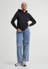 Sweatshirt à capuche noir associé à un jean large déchiré bleu clair et des sneakers de sport gris. Le mannequin pose avec une main sur la hanche.
