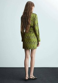 Grün gemustertes Kleid mit langen Ärmeln und figurbetontem Schnitt. Verfügt über geschwungene, abstrakte Designs in dunkleren Farbtönen. Getragen mit schwarzen flachen Schuhen.