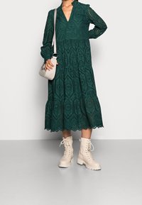 Grön spetklänning med långa ärmar, V-hals och flera lager i kjolen. Kombinerad med krämfärgade kängor och en ljusgrå axelväska.