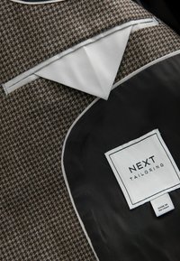 Brown houndstooth patterned fabric with light gray trim, featuring a black interior pocket labeled "NEXT TAILORING" and care tag.