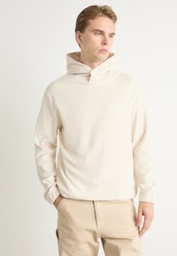 Sudadera de canalé color crema con cuello de embudo, que presenta un corte relajado y mangas largas, combinada con pantalones de un beige claro.