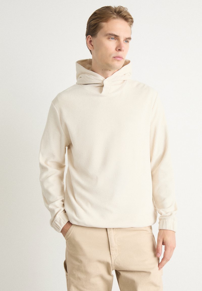 Sudadera de canalé color crema con cuello de embudo, que presenta un corte relajado y mangas largas, combinada con pantalones de un beige claro.