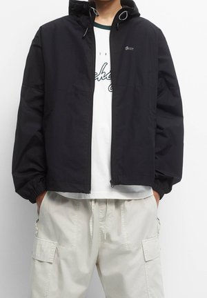 Osoba nosząca czarną kurtkę z kapturem na białej koszulce z nadrukiem oraz jasne spodnie typu cargo, z rękami w kieszeniach.