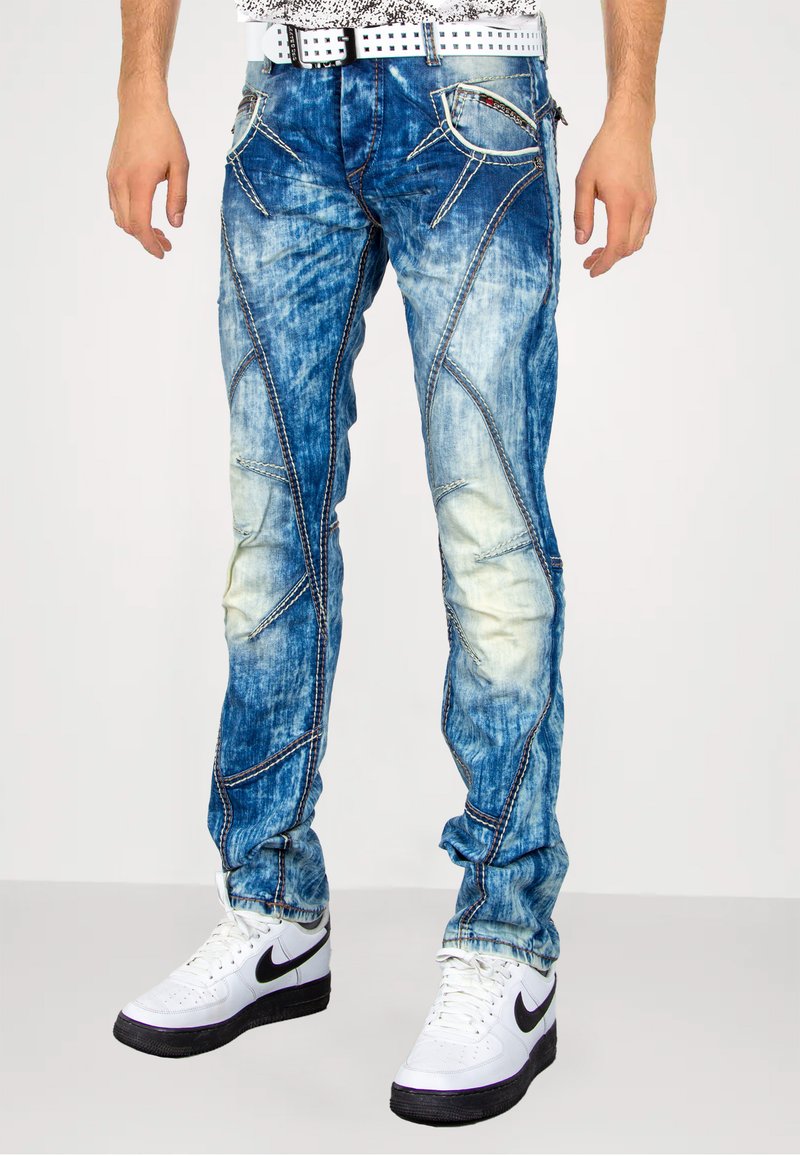 Blauwe denim jeans met een versleten afwerking, contrasterende stiksels, uniek paneelontwerp en vijf zakken, gedragen met wit- en zwartkleurige sneakers.