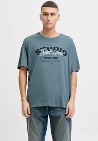 Grå bomulds t-shirt med korte ærmer, der har sorte og hvide grafikker, der står "STUDIO Jack & Jones" og "SINCE 1990."