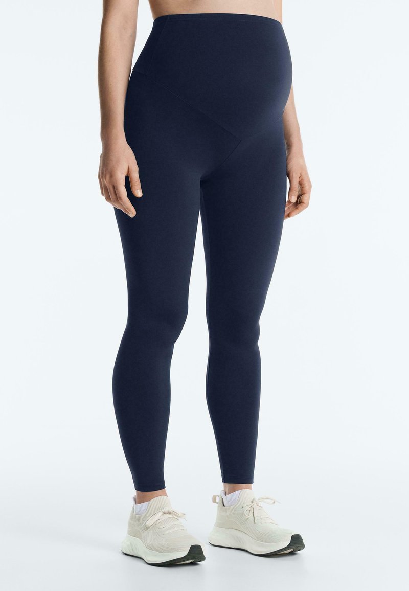 Personne enceinte portant des leggings de maternité bleu marine et des chaussures de sport beige, debout devant un fond blanc uni.