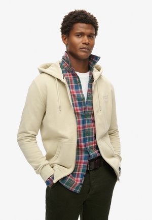 Felpa beige con zip e coulisse, indossata sopra una camicia a quadri multicolore. Presenta una tasca frontale e una cintura scura, su uno sfondo bianco.