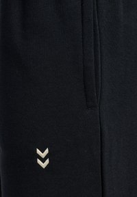 Svarta mjukisbyxor med mjuk textur, resårmidja, sidofickor och en krämfärgad logotyp med två nedåtriktade v-former.