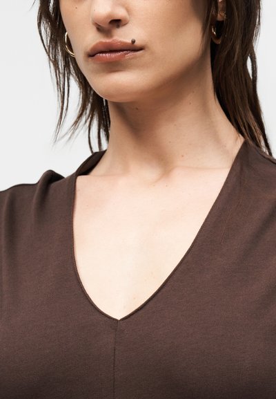Κοντινό πλάνο ενός ατόμου που φοράει καφέ τοπ με V-λαιμόκοψη, με ανοιχτόχρωμο δέρμα, κοντά σκούρα μαλλιά, ελιά πάνω από το επάνω χείλος και κρίκο στο αυτί.