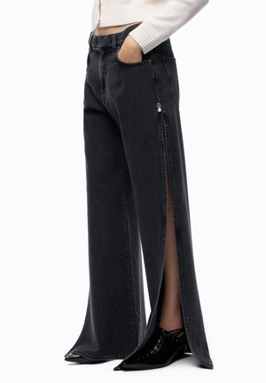 Vaqueros negros de pierna ancha con aperturas laterales altas y pequeño detalle plateado, combinados con zapatos negros puntiagudos y un jersey corto blanco.
