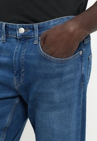 Jeans de denim azul com corte reto, apresentando um bolso visível, rebite metálico e detalhes de costura ao longo das costuras e da cintura.