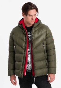 Olivgrön pufferjacka med röd huva och dragkedjor. Den har en chevronquiltad design och en slät, glansig yta.