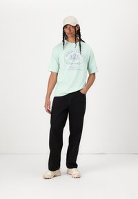 Mint green t-shirt with a graphic logo, paired with black loose-fitting pants and light sneakers. The model is wearing a beige cap.