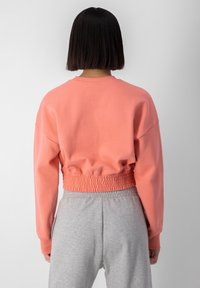 Kvinna med rakt, mörkt hår sedd bakifrån, iförd en korallfärgad kort sweatshirt med elastisk nederkant och ljusgråa sweatpants.