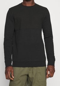 Svart sweatshirt med ett strukturerat övre panel och en slät nedre del, med långa ärmar och rundad halsringning. Tillverkad av mjukt tyg.