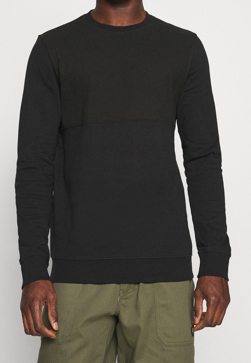 Svart sweatshirt med ett strukturerat övre panel och en slät nedre del, med långa ärmar och rundad halsringning. Tillverkad av mjukt tyg.