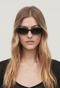 Gafas de sol negras, rectangulares, con montura de plástico grueso y lentes oscuros. Presentan una textura suave y un diseño minimalista.