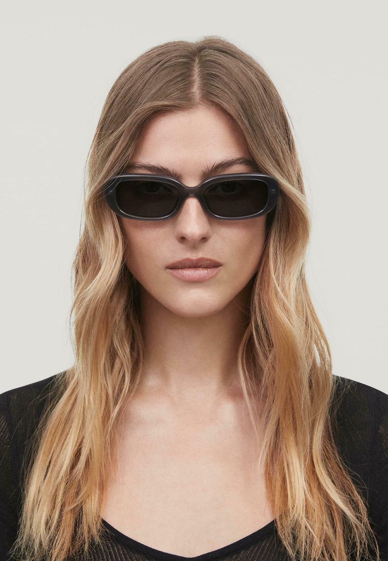 Gafas de sol negras, rectangulares, con montura de plástico grueso y lentes oscuros. Presentan una textura suave y un diseño minimalista.