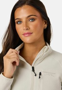 Light beige fleece jacket with a zip-up collar, featuring black drawstrings and embroidered "Swedemount" logo on the chest. Soft texture.