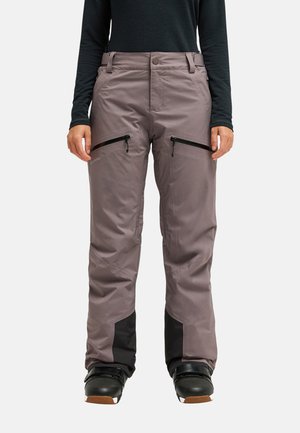 Person trägt graue Outdoor-Hose mit schwarzen verstärkten Bündchen, Reißverschlusstaschen am Oberschenkel und schwarzen Stiefeln, steht vor einem schlichten Hintergrund.