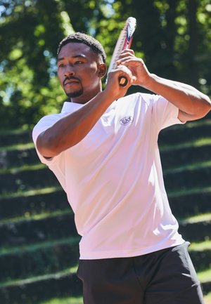 Polo shirt bianco con logo, pantaloni corti neri, tiene una racchetta da tennis, sullo sfondo verde con gradini di pietra a terrazze.