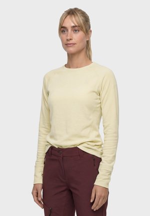 Frau mit blondem Haar, die ein hellgelbes Langarmshirt und dunkelrote Hosen trägt, steht vor einem einfarbigen Hintergrund.