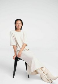 Strik creme kjole med en afslappet pasform og korte ærmer, parret med knæhøje beige støvler, siddende på en sort stol. Blødt, tekstureret stof.