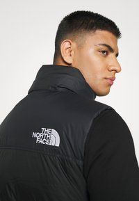 Black insulated vest with a high collar, featuring a white logo on the left side; smooth texture and a sleek design.