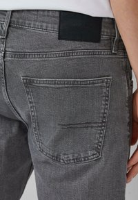 Close-up van een persoon die grijze denim jeans draagt, waarbij de achterzak en het zwarte leren merklabel op de tailleband zichtbaar zijn.