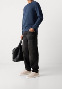 Uomo che indossa una felpa blu navy, jeans neri larghi, sneaker beige e tiene in mano una borsa da viaggio in pelle nera con la mano sinistra.