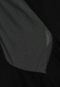 Melna apģērba detaļa ar caurspīdīgu acu paneļu, uz kura ir mazi punkti, mīkstu tekstūru un kontrastējošiem gludiem auduma malām.