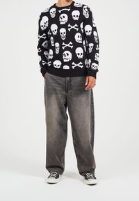 Svart stickad tröja med vita dödskalle- och korsbenmönster; matchad med grå, löst sittande jeans och svartvita sneakers.