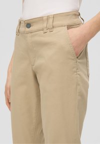 Personne portant un pantalon beige, la main dans la poche droite, avec un haut blanc côtelé visible à la taille, sur un fond clair uni.