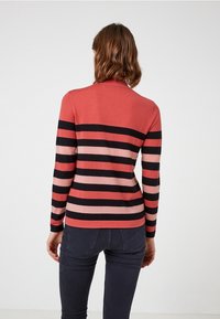 Femme portant un pull à manches longues avec des rayures horizontales noires, corail et roses, associée à un jean ajusté foncé, tournée de dos à la caméra.