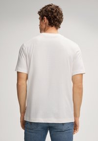 Un homme aux cheveux bouclés se tient de dos, portant un t-shirt blanc à manches courtes et un jean bleu, devant un fond uni.