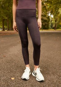 Leggings i mörklila med leopardmönster och kontrasterande sömmar, i kombination med vita sportskor på en grusväg.