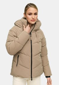 Vrouw in een beige gewatteerde jas met een hoge kraag en capuchon, die met één hand de kraag vasthoudt, tegen een effen achtergrond.