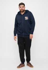 Navy zip-up hoodie with a logo print on the chest, paired with black cargo pants and brown sneakers. Hoodie features a drawstring and kangaroo pockets.