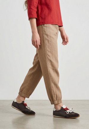 Persoon draagt beige joggingsbroek en zwarte sneakers met rode en witte veters en loopt over een gladde vloer tegen een effen achtergrond.