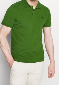 Homme portant un polo vert à manches courtes avec un logo brodé, associé à un pantalon clair, main dans la poche, sur un fond blanc uni.