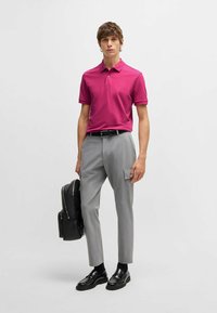 En figursydd magenta pikétröja, grå skräddarsydd byxor med sidfickor, svart bälte och skor, hållande en svart ryggsäck. Slät tyg och rena linjer.