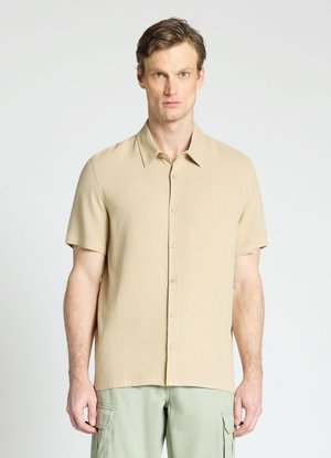 Un homme se tient face à l'avant, portant une chemise beige à manches courtes boutonnée et un pantalon cargo vert clair sur un fond uni.