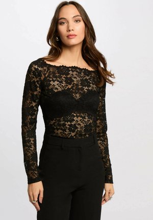 Donna con capelli castani lunghi che indossa una maglia nera a maniche lunghe con pizzo floreale e pantaloni neri a vita alta, su uno sfondo chiaro e uniforme.