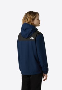 Granatowa kurtka z czarną górną częścią i kapturem. Posiada elastyczne mankiety i logo na plecach. Gładka tekstura tkaniny.