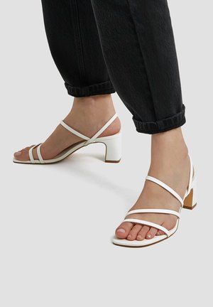 Sandales à talons blancs avec brides, dotées d'un talon carré, présentant des lanières fines et une texture lisse. La hauteur du talon est modérée, convenant à une tenue décontractée.