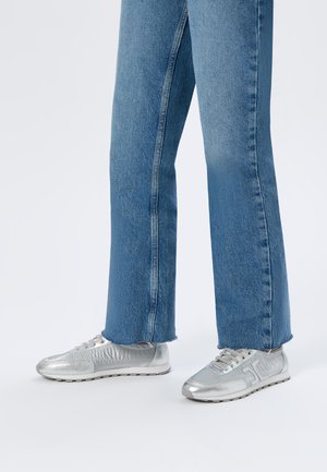Jambes portant un jean en denim bleu avec des ourlets bruts et des baskets basses métalliques argentées sur un fond clair uni.