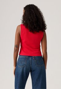 Débardeur rouge à côtes avec un design ajusté, présentant de larges emmanchures et une longueur cropped, associé à un pantalon en denim bleu clair.